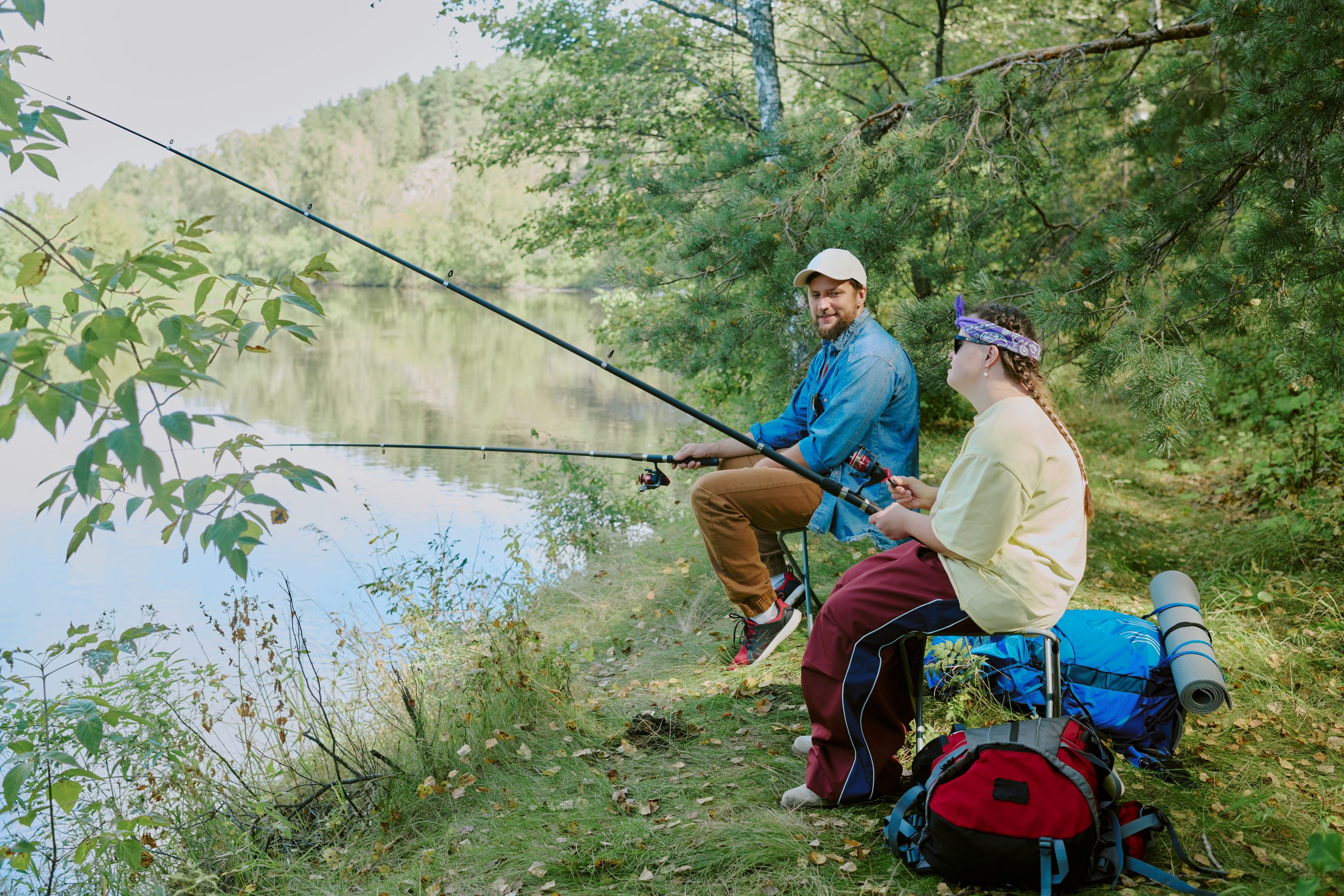 Young man and young woman who has Down syndrome sit lakeside with fishing poles, talking.